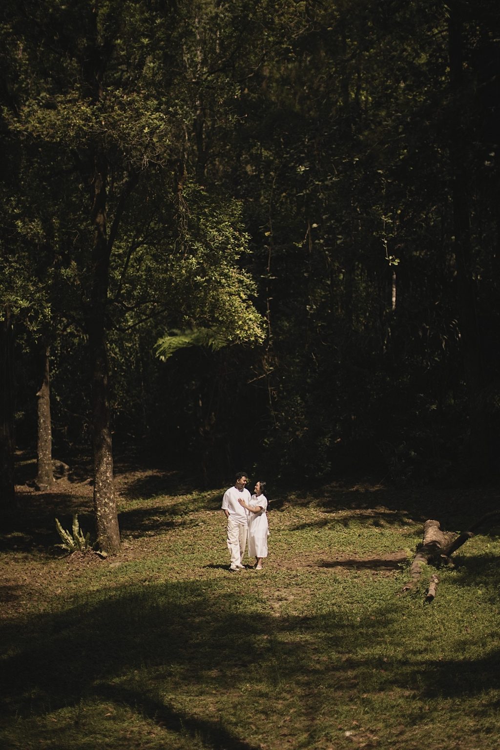Brian and Sasa in white standing close together in a sunlit forest clearing