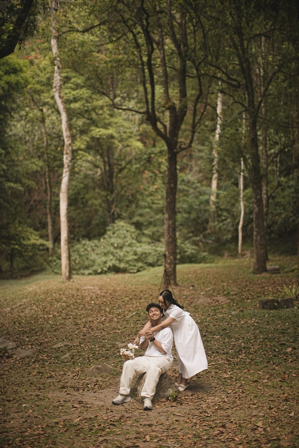 Brian sitting on a rock as Sasa embraces him from behind beneath tall trees