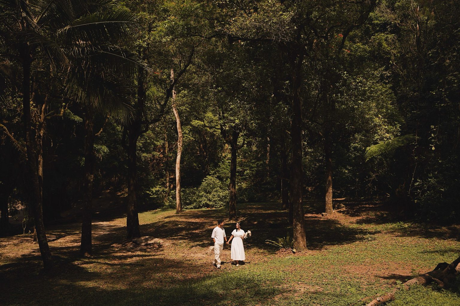 Brian and Sasa walking hand in hand across a sunlit clearing as Sasa holds a bouquet